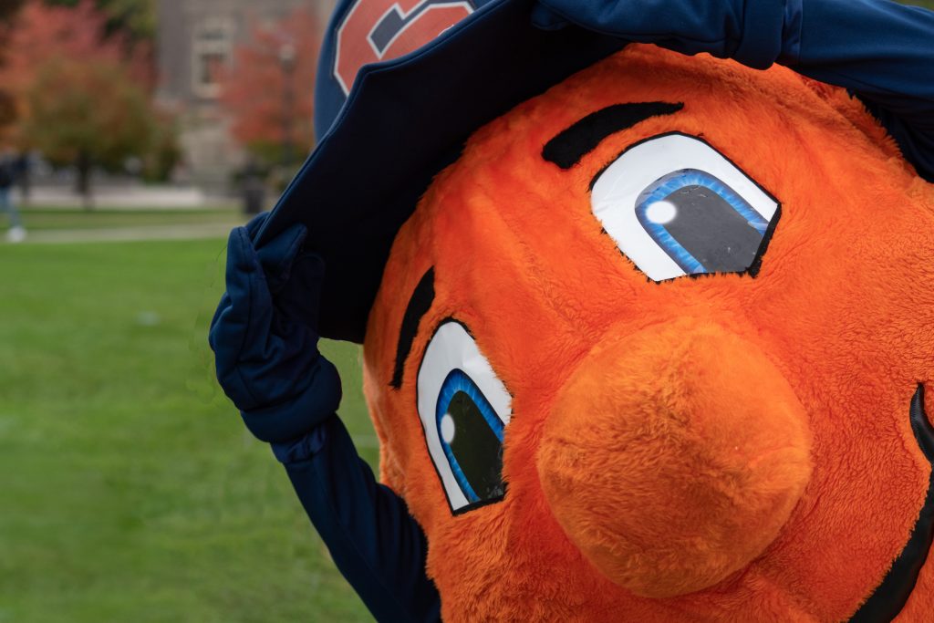 Mascot Otto the Orange poses with a large baseball cap that has a Syracuse University logo on it.