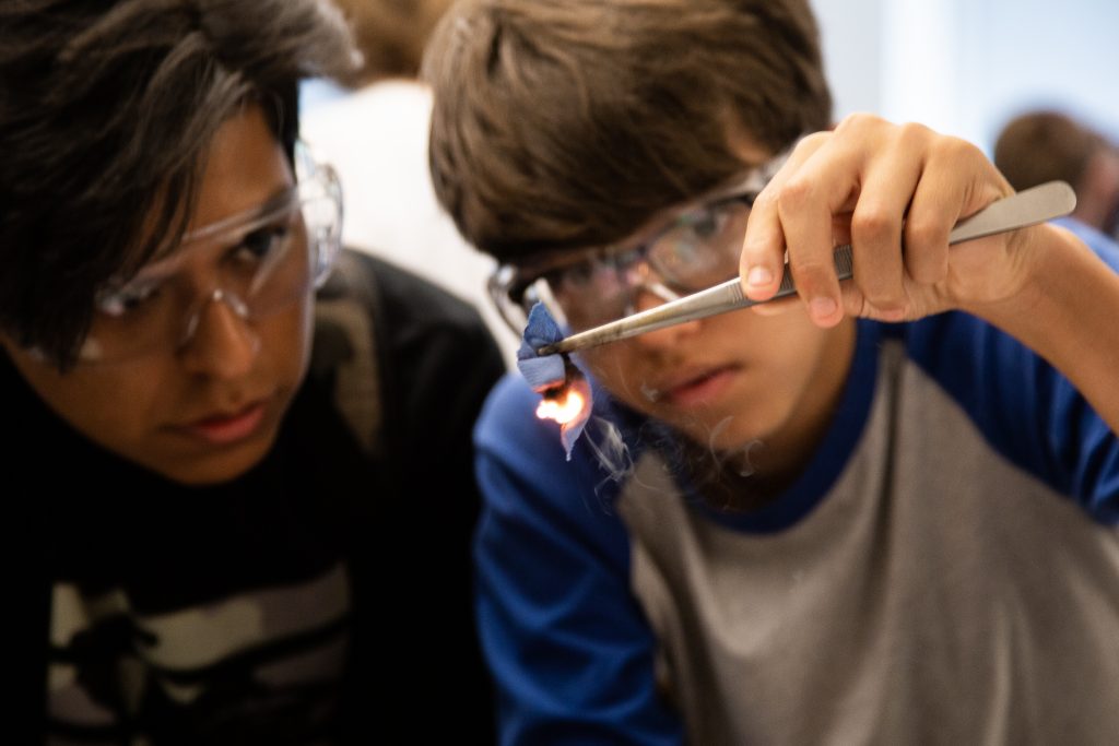 Two students wearing safety goggles look closely at an experiment that includes a small flame.