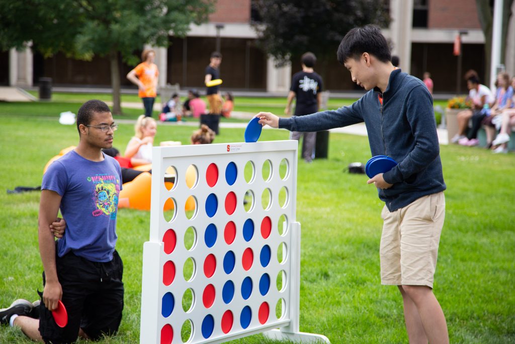 Two students sit in the grass, playing Connect Four