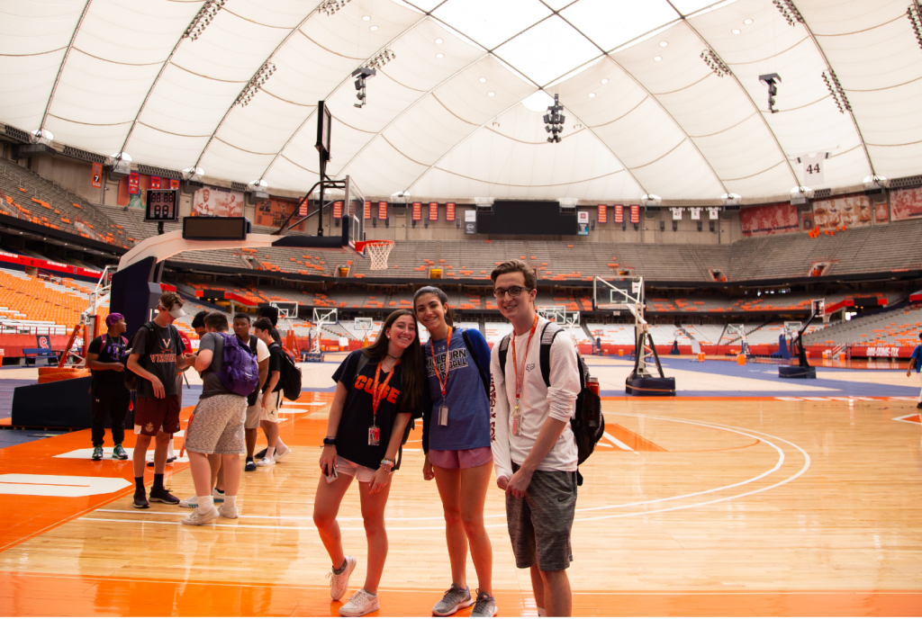 Three students wearing Syracuse University shirts and lanyards stand on the basketball court