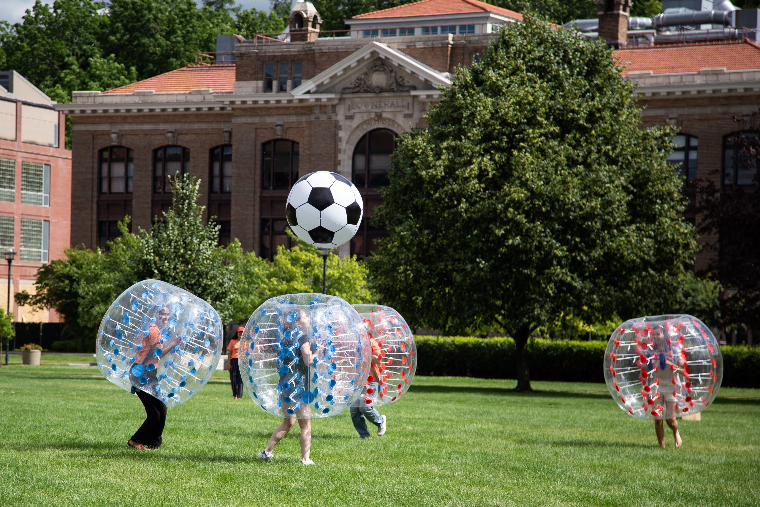 Several students wearing giant plastic bubbles, hitting an inflated soccer ball around outside.